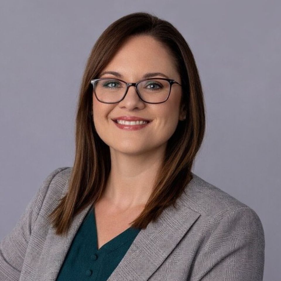 A woman with straight brown hair and glasses smiles at the camera. She is wearing a gray blazer over a dark green top, with a light purple background behind her.