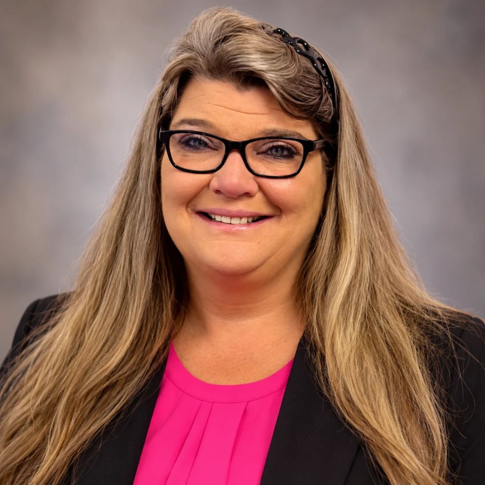A smiling woman with long blonde hair, wearing black-rimmed glasses and a black blazer over a bright pink top.